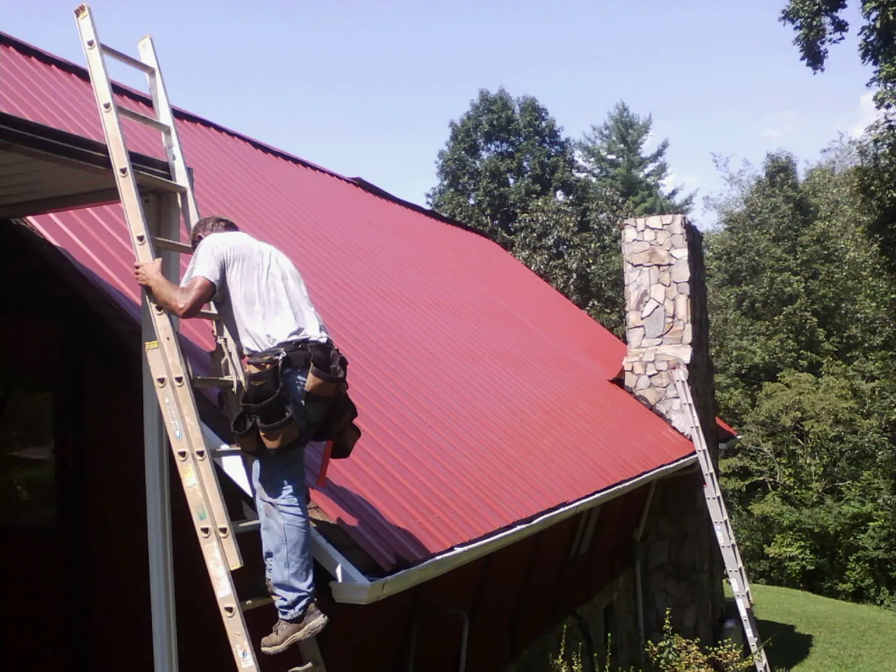Roofer on a ladder beside a red metal roof and stone chimney — Soffit & Fascia Repair by Roofing Brothers in Denmark